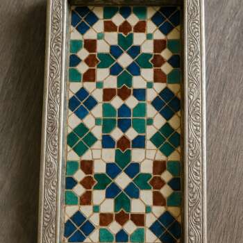 Support de stylo rectangulaire en zellige marocain, avec un cadre argenté orné de motifs floraux et une mosaïque colorée de carreaux bleus, verts, marrons et blancs. Posé sur un bureau en bois clair, il mêle artisanat traditionnel et design contemporain.
